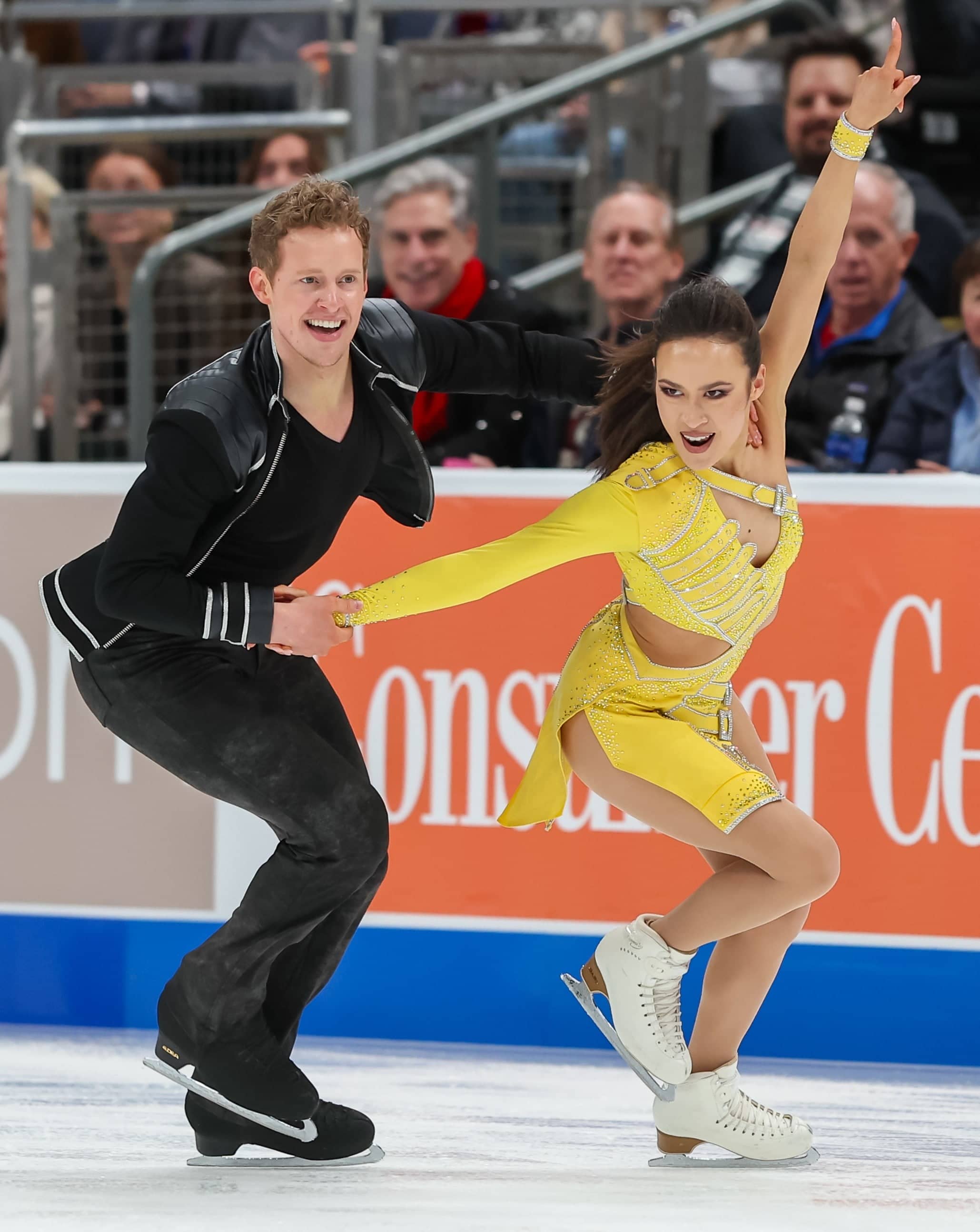 Madison Chock and Evan Bates compete together. Madison is a woman with long black hair. She is wearing a yellow long sleeve dress. She is pointing to the sky with her left hand and holding Evan's hand with her right. Evan is a man with blonde curly hair wearing a black top with a black jacket and black pants